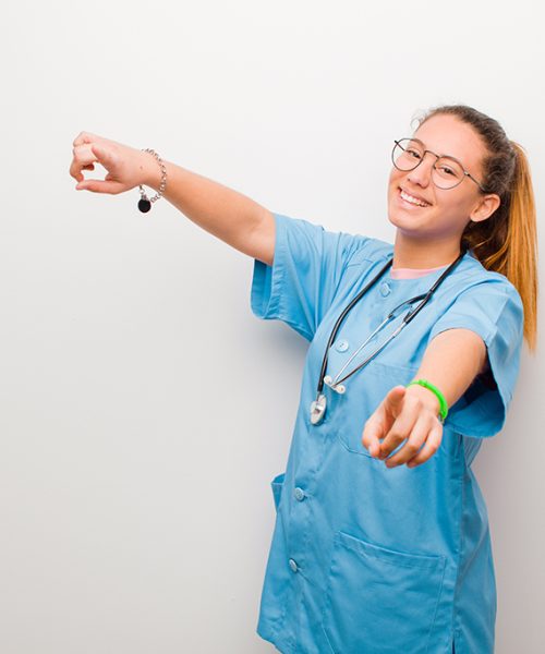 young latin nurse feeling happy and confident, pointing to camera with both hands and laughing, choosing you against white wall